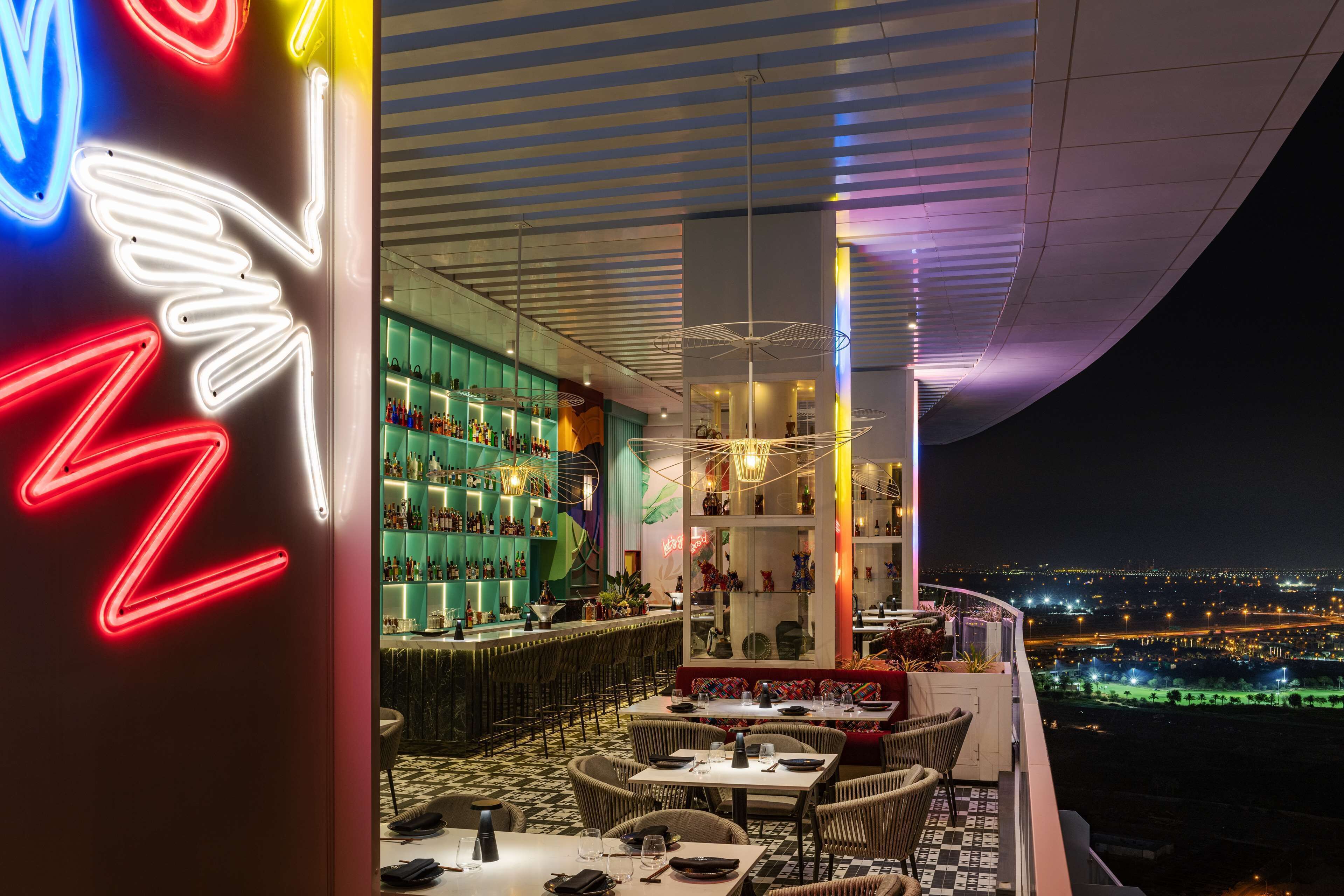 A rooftop restaurant with a view of the cityscape at night, featuring neon signage and modern interior design.