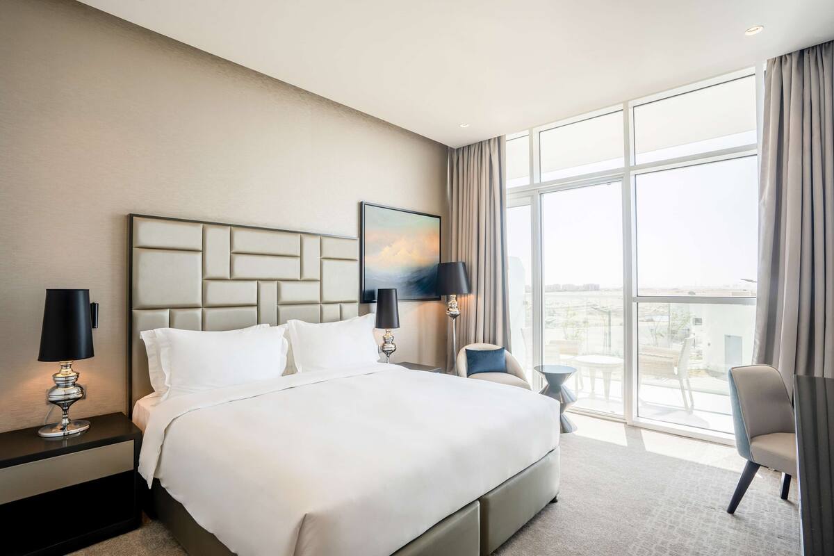 A modern hotel room with a large bed, two bedside lamps, a nightstand, a chair, and a large window offering a city view.