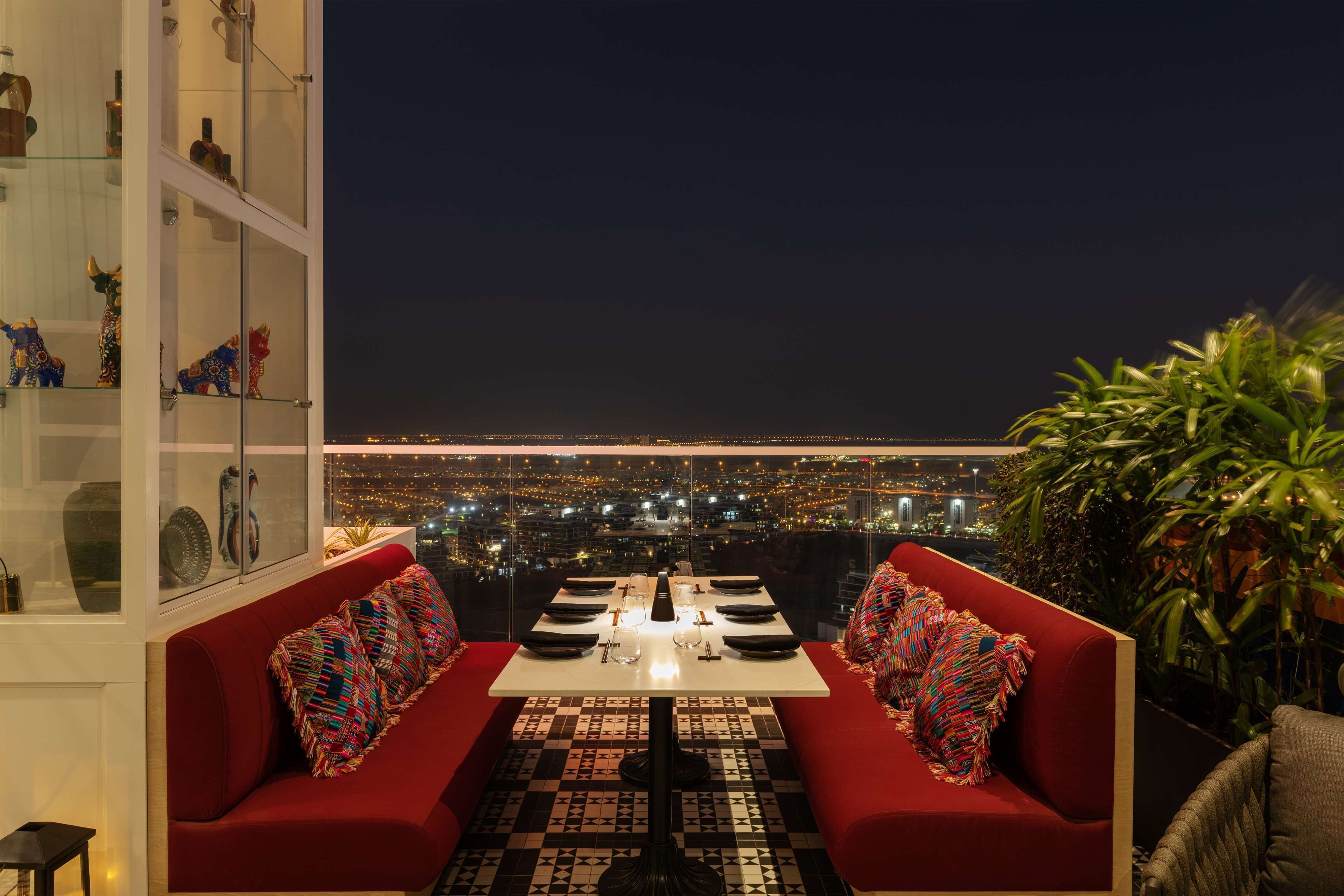 Un comedor con una mesa dispuesta para dos personas, un sofá rojo y vistas nocturnas de la ciudad.