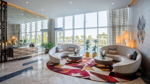 A modern lobby with a curved sofa set, a patterned rug, and large windows offering a view of palm trees. - Radisson Dubai Damac Hills (Dubai)