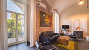 A living room with a black leather sofa, a wooden coffee table, and a TV. There is a large window with curtains, a brick wall with an abstract painting, and a small table with a mirror and a chair. - THE BELLINI HOUSE (Naples)