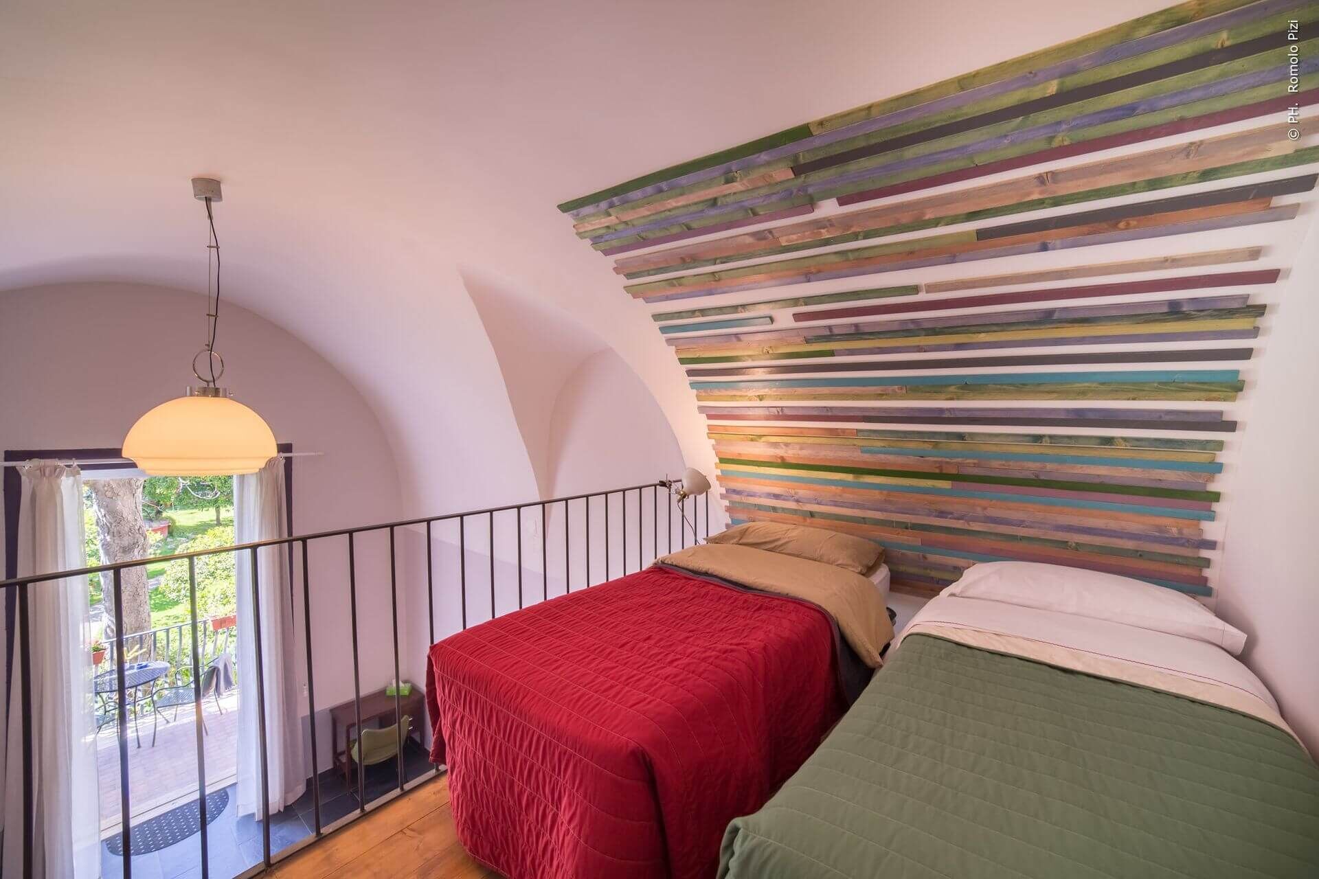 A bedroom with a sloped ceiling, a bed with a red and green quilt, a colorful wooden headboard, and a hanging light fixture.