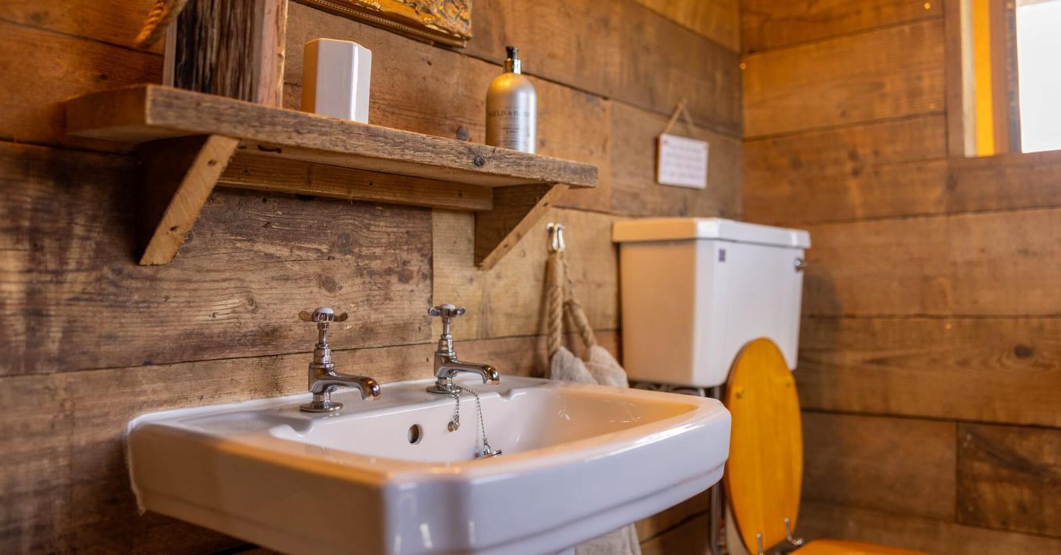 A rustic bathroom with a wooden wall, a white sink, two faucets, a wooden shelf with toiletries, and a toilet.