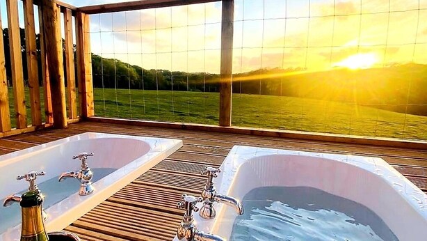 A wooden deck with two hot tubs and a view of the sunset over a body of water. - Luxury Safari Lodge-sleeps 4/6-deer Park-dogs Ok (Crediton)