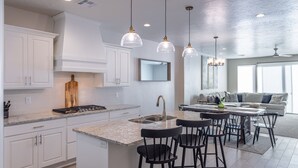 A modern kitchen with white cabinets, a central island with bar stools, pendant lighting, and a dining area with a sofa and chairs.
