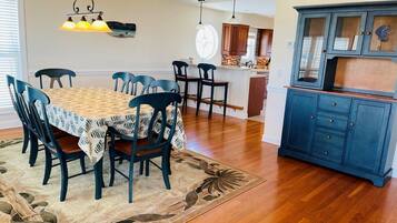 A dining area with a table, chairs, a rug, a hanging light fixture, and a kitchen area with cabinets.