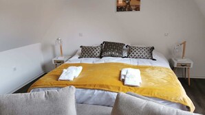 Un lit bien fait, recouvert d’une couverture jaune, avec deux oreillers et deux serviettes pliées. Il y a deux tables de chevet avec des lampes et un petit tableau accroché au mur, au-dessus du lit.