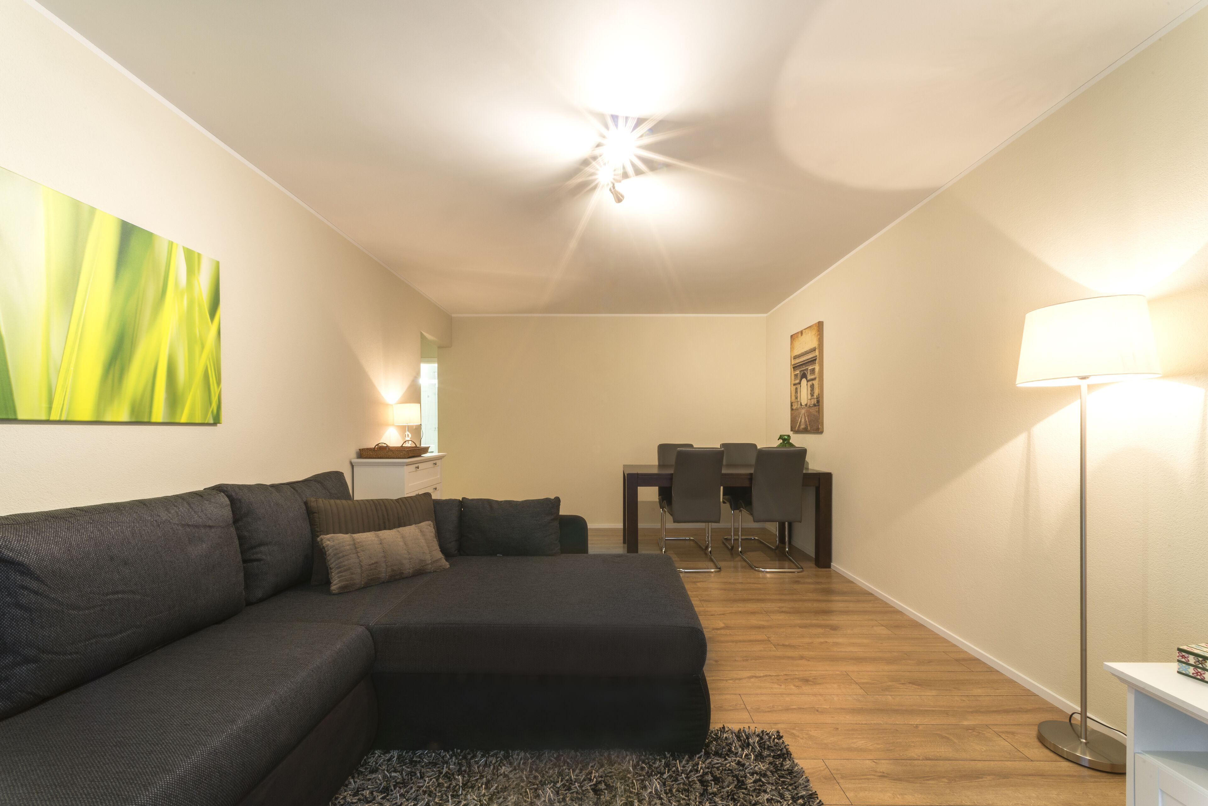 A modern living room with a sectional sofa, a dining table with chairs, a lamp, and a painting on the wall.