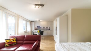 A modern room with a red sofa, a white bed, and a kitchen in the background. - RELOC Serviced Apartments Wallisellen 36 (Wallisellen)