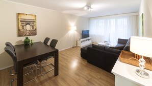A modern living room with a dining area, featuring a sofa set, a dining table with chairs, a TV, and a lamp. - RELOC Serviced Apartments Wallisellen 36 (Wallisellen)