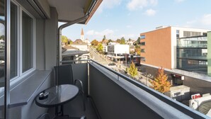 A balcony with a table and chairs, overlooking a street with buildings and a church steeple. - RELOC Serviced Apartments Wallisellen 36 (Wallisellen)