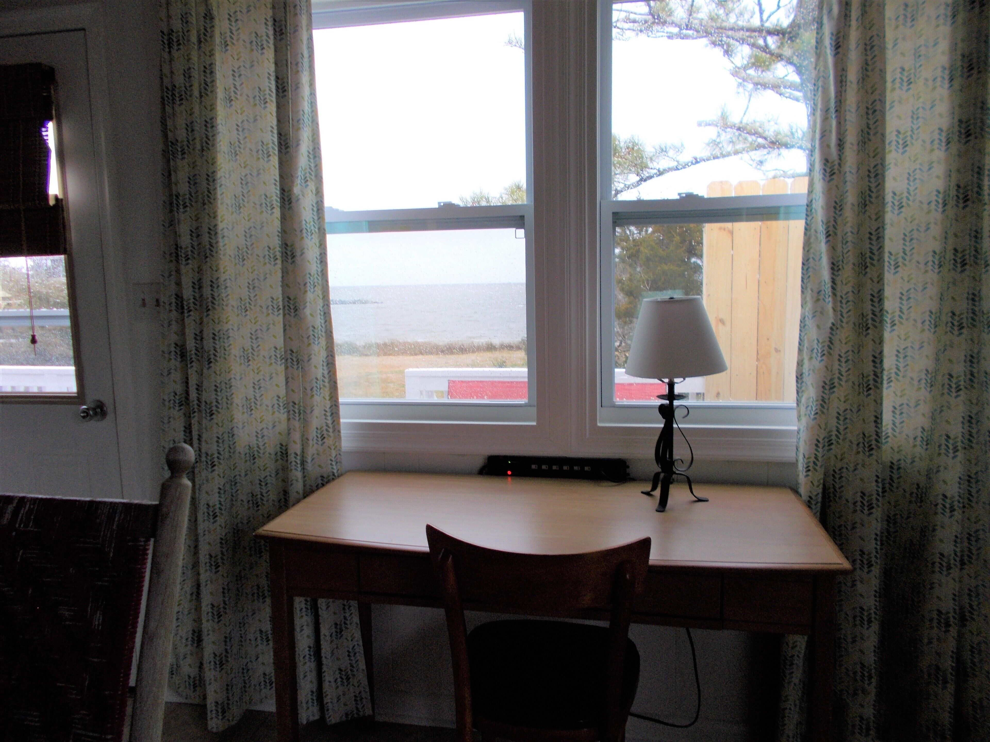 A room with a desk, chair, and a lamp. There is a window with curtains, and a view of the outside.