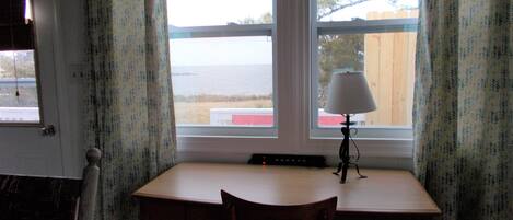 A room with a desk, chair, and a lamp. There is a window with curtains, and a view of the outside.