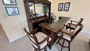 A dining area with a long wooden table, four upholstered chairs, a bar counter with a mirror, and two framed pictures on the wall.