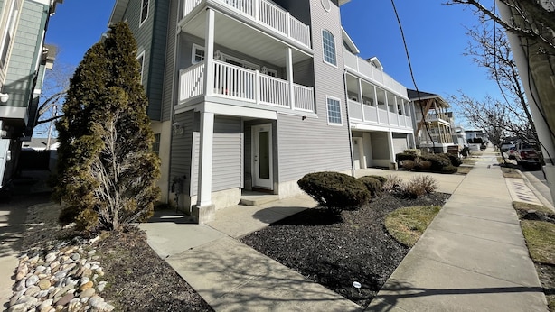 A two-story residential building with a balcony on the second floor, a garage, and a well-maintained front yard with a stone border and shrubs.