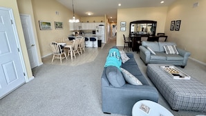 A spacious living area with a grey sofa, a patterned ottoman, and a dining table with chairs. A ceiling fan and recessed lighting are visible.