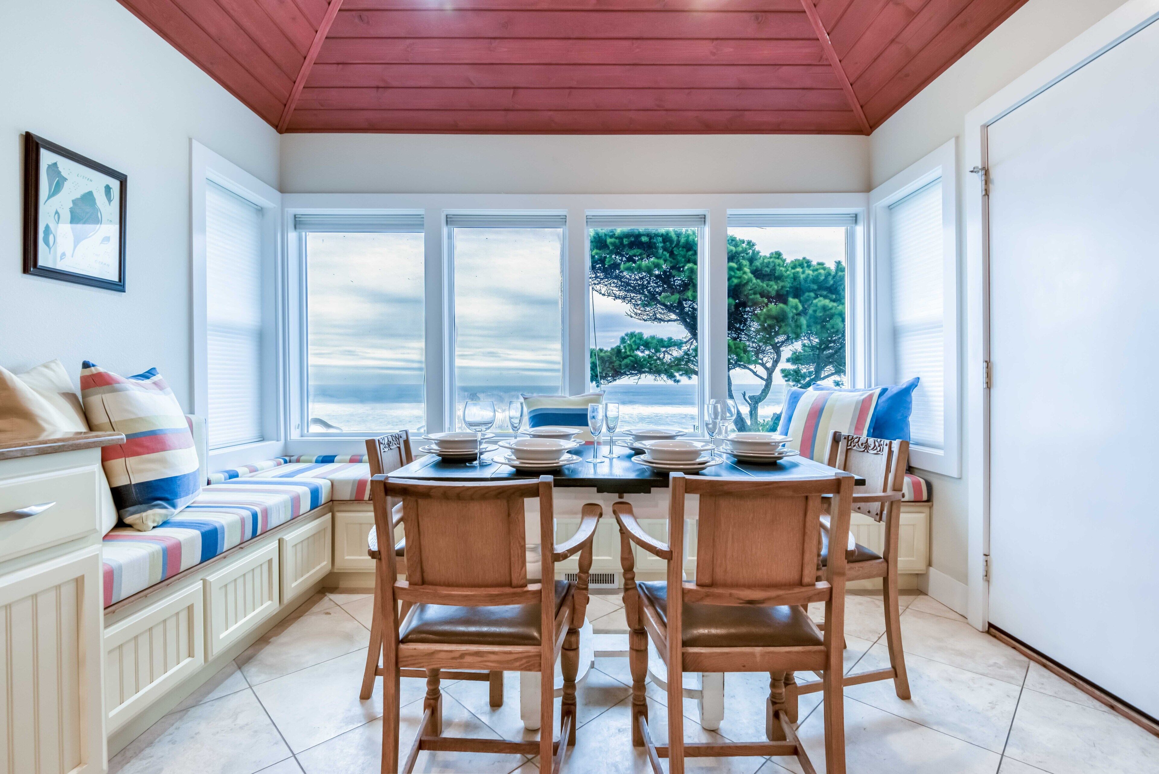 Un coin repas avec une table dressée pour quatre personnes, des chaises en bois, un banc avec des coussins et de grandes fenêtres offrant une vue sur les arbres et le ciel.