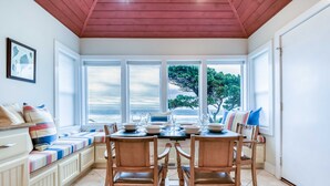 A dining area with a table set for four, wooden chairs, a bench with cushions, and large windows offering a view of trees and the sky.