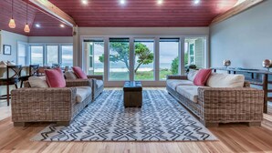 A spacious living room with wicker furniture, a patterned rug, and large windows offering a view of the outdoors.