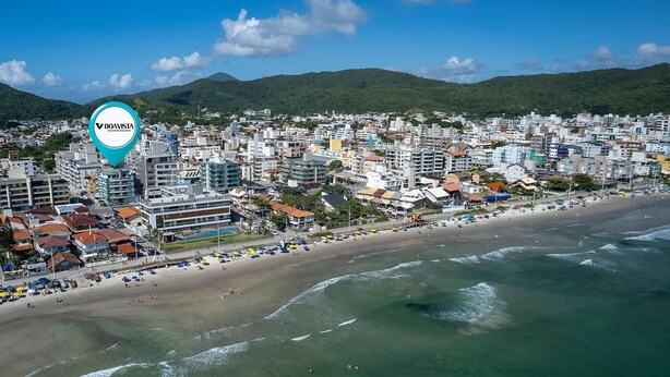 A coastal cityscape with a beach, buildings, and mountains in the background. - Zancanaro Apto 202  641D (Bombinhas)