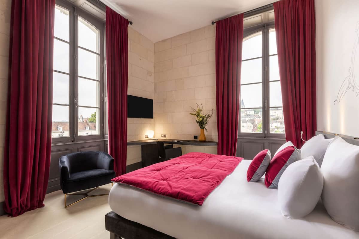 A hotel room with a large bed, red curtains, a black armchair, a desk, and a view of the city through large windows.