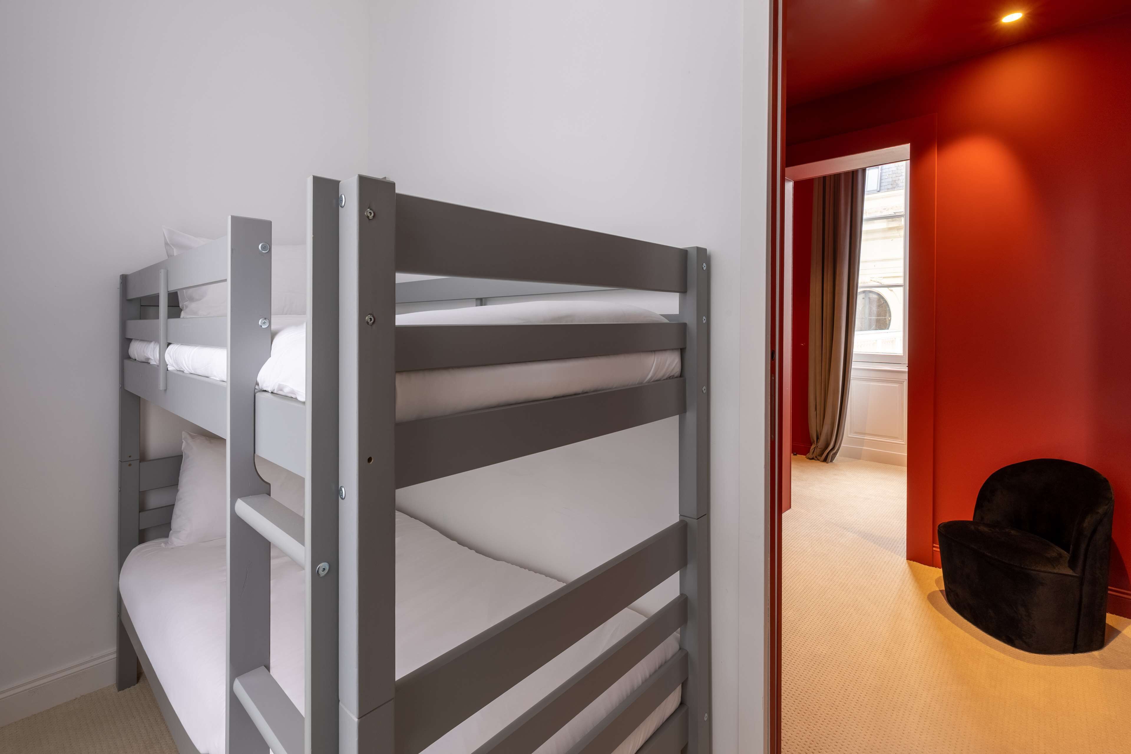 A bunk bed with grey metal frames and white bedding, a black chair, and a red wall in the background.