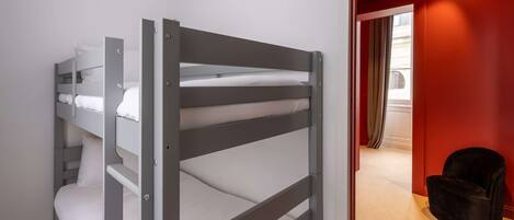 A bunk bed with grey metal frames and white bedding, a black chair, and a red wall in the background.