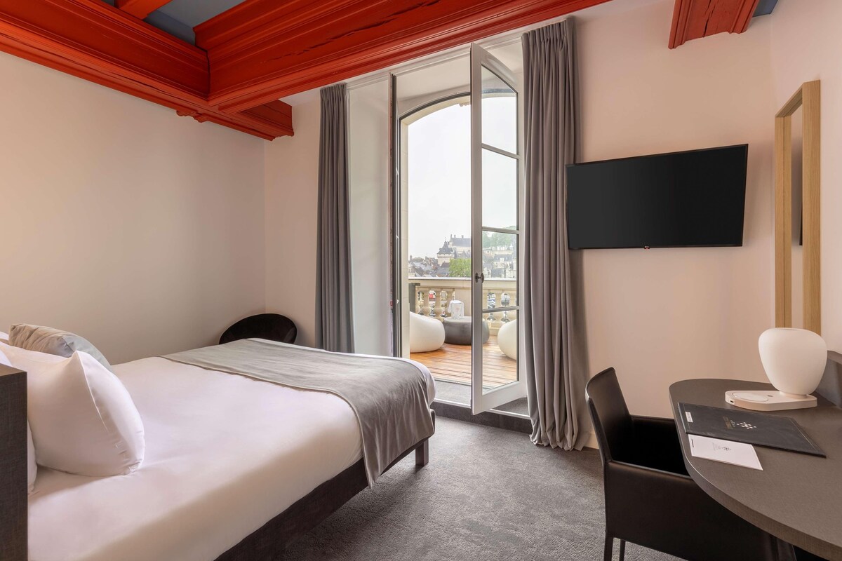 A hotel room with a bed, a desk, a chair, a TV, and a balcony with curtains.