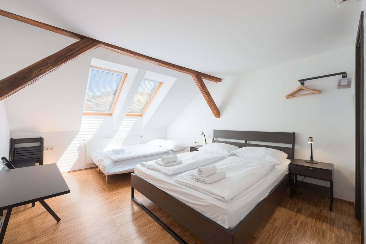 A modern bedroom with a sloped ceiling, two beds, a desk, and a hanging light fixture.