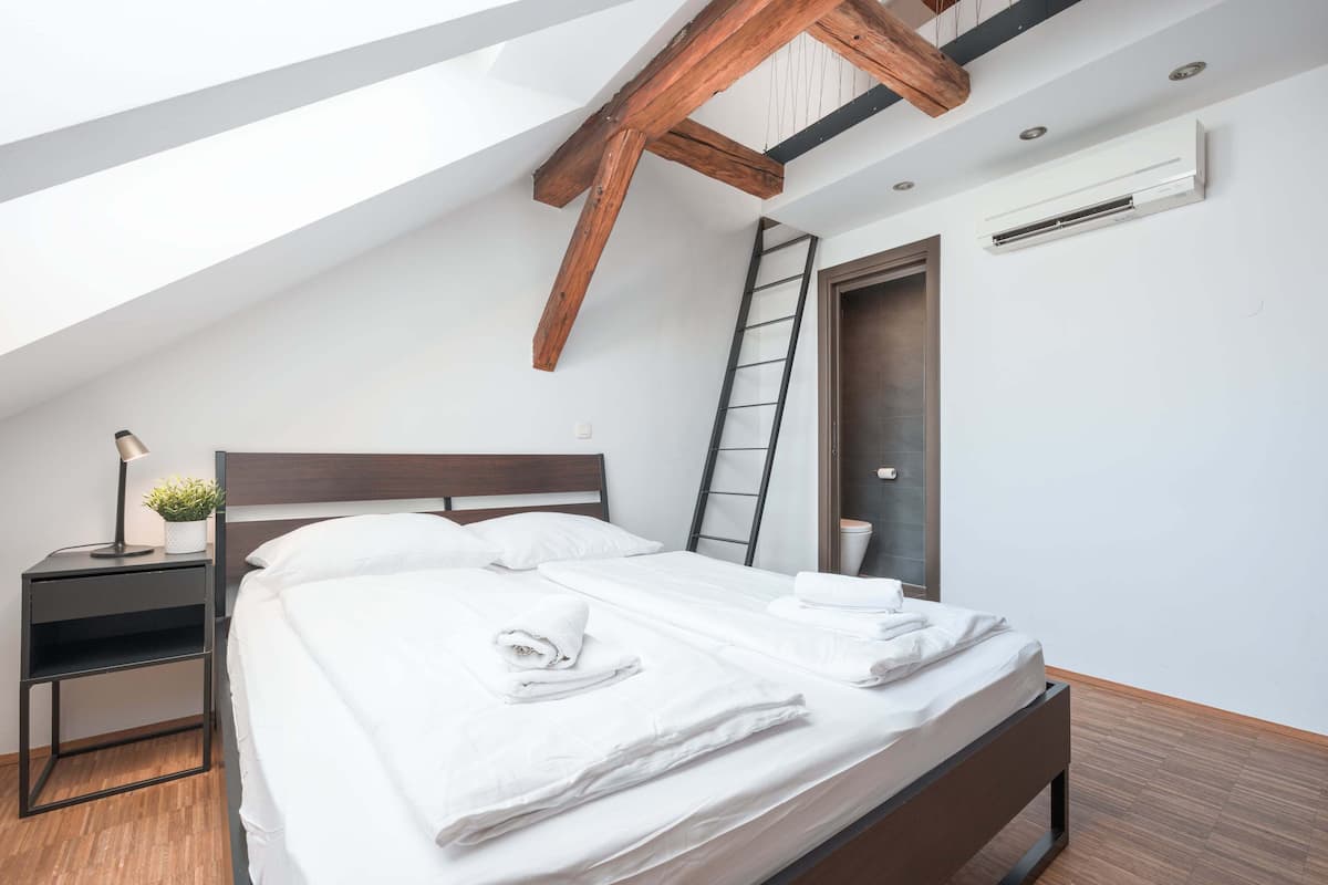 A modern bedroom with a sloped ceiling, a wooden headboard, a bedside table with a lamp, and a neatly made bed.