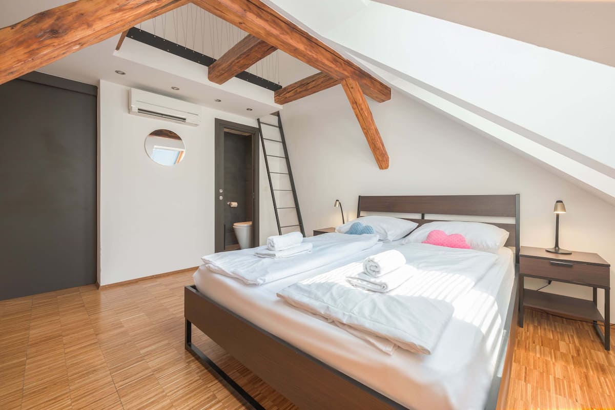 A bedroom with a sloped ceiling, wooden beams, a bed with white linens, a bedside table, and a nightstand.