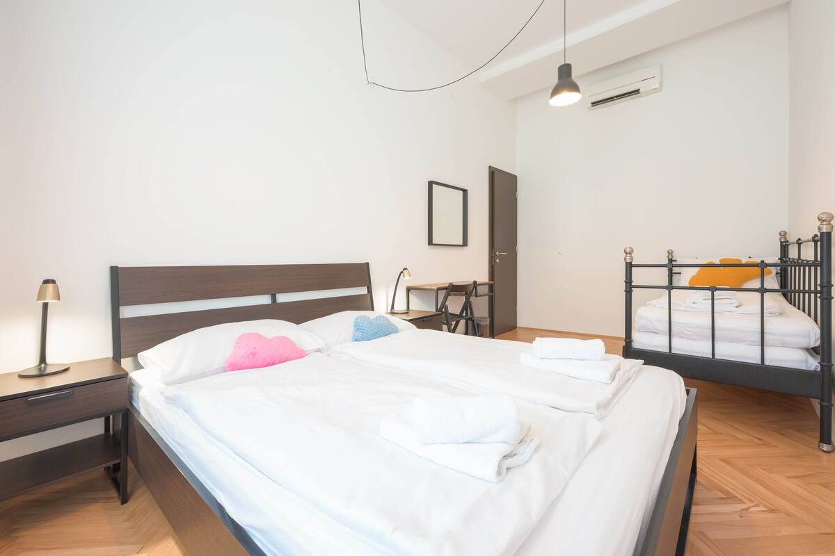 A hotel room with a large bed, a desk, a chair, a wardrobe, and a small table.