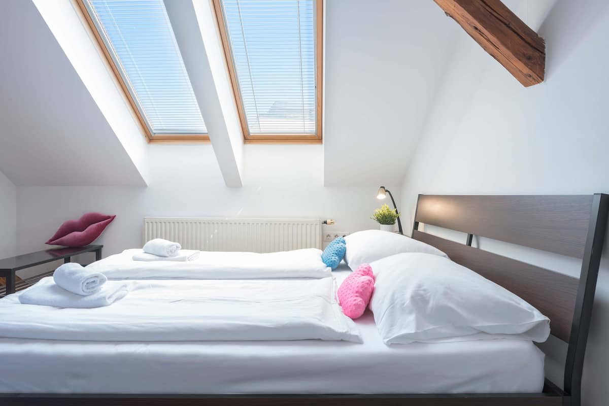 A bedroom with two beds, skylights, a radiator, a nightstand, a lamp, and a chair.
