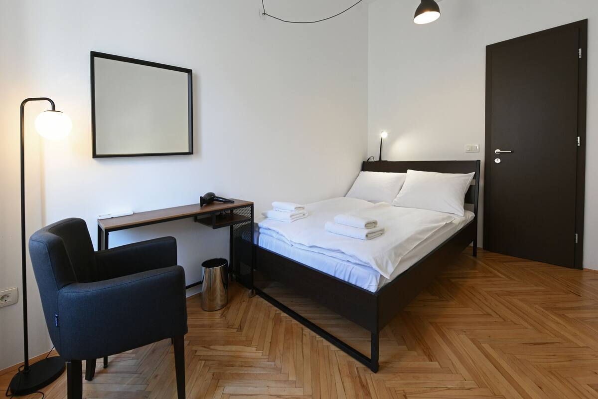 A modern hotel room with a wooden herringbone floor, a bed with white linens, a desk with a chair, a standing lamp, and a dark door.