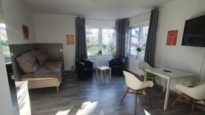 A modern living room with a sofa, a dining table, and a TV. There are curtains on the windows, and a plant is visible on the windowsill. - Krabbenkieker (Norden)
