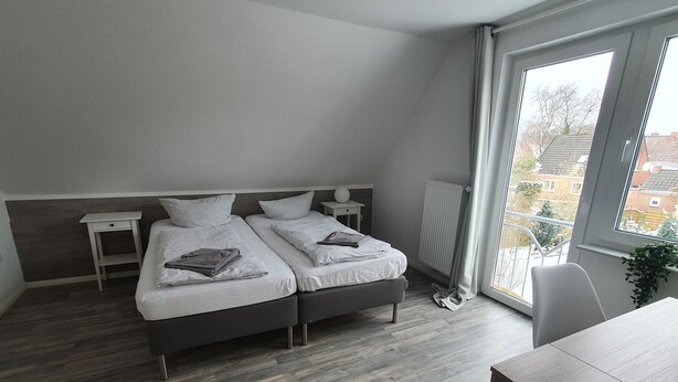 A modern bedroom with two beds, a small table, and a large window with a view of buildings. - Krabbenkieker (Norden)