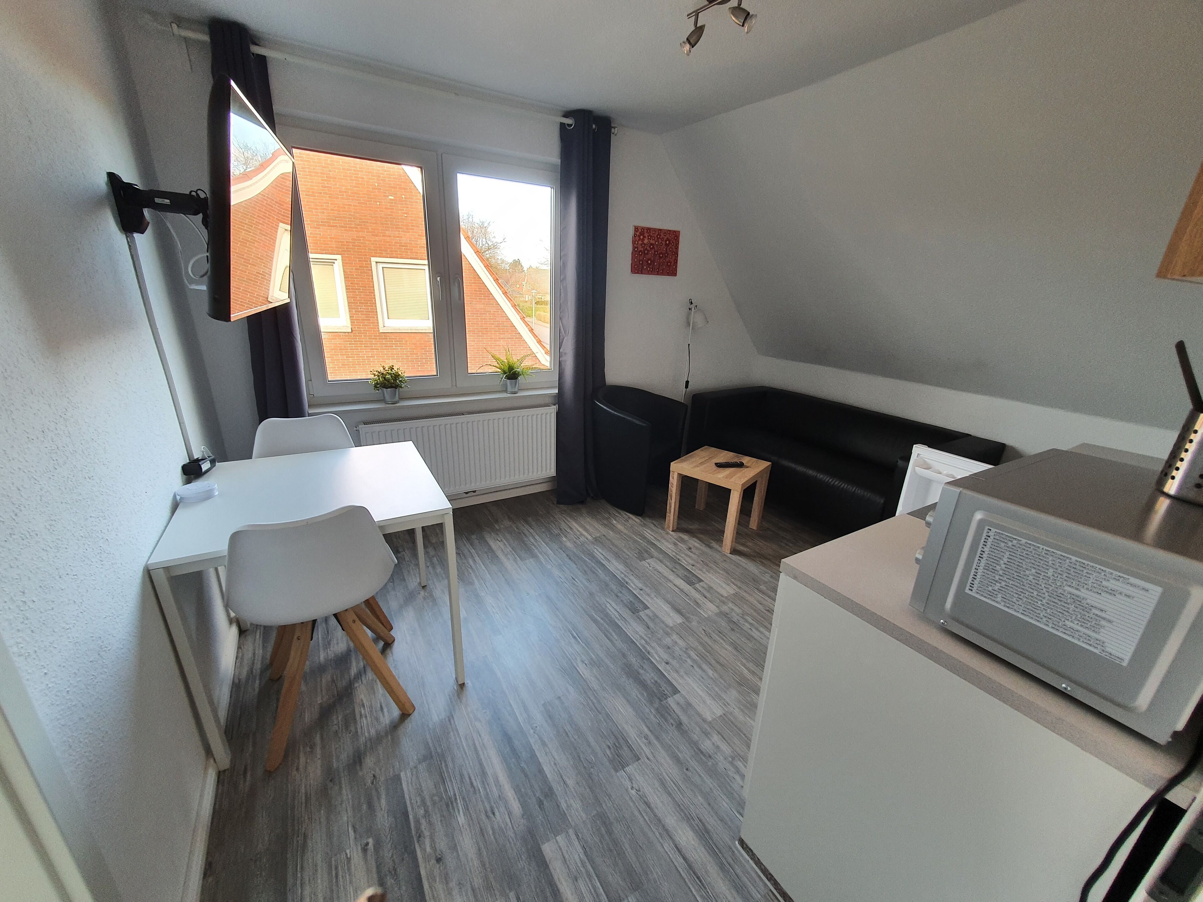 Habitación pequeña con escritorio blanco, sofá negro, taburete de madera, radiador blanco y ventana con vista a edificios.