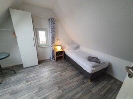 A small, neatly arranged bedroom with a single bed, a bedside table with a lamp, a chair, and a window with curtains.