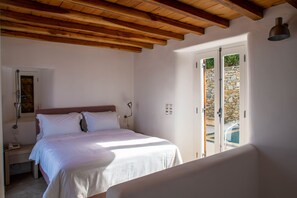 A bedroom with a wooden ceiling, a bed with white bedding, a nightstand, a lamp, and a door leading to an outdoor area. - Villa Ester with Heated Pool & Hot Tub (Mykonos)