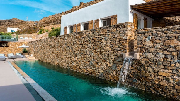 Une piscine aux murs de pierre, agrémentée d’une cascade, entourée d’un bâtiment blanc avec des portes et des fenêtres en bois.