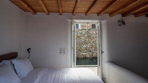 Une chambre avec un plafond en bois, un lit avec une literie blanche, un mur en pierre à l’extérieur et une petite fenêtre.