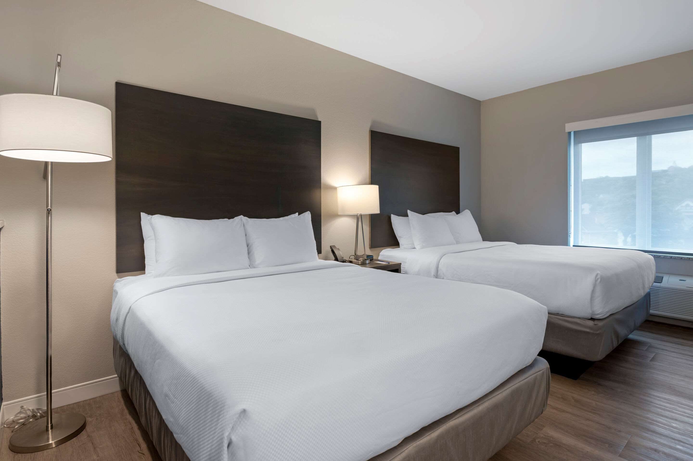 A hotel room with two beds, a floor lamp, a bedside table with a lamp, and a large window.