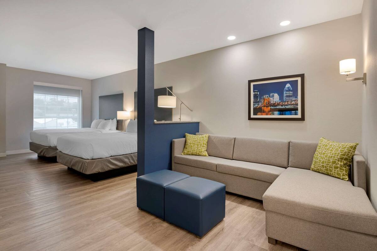 A hotel room with a bed, two bedside tables, a sofa, an ottoman, a large window, and a cityscape painting on the wall.