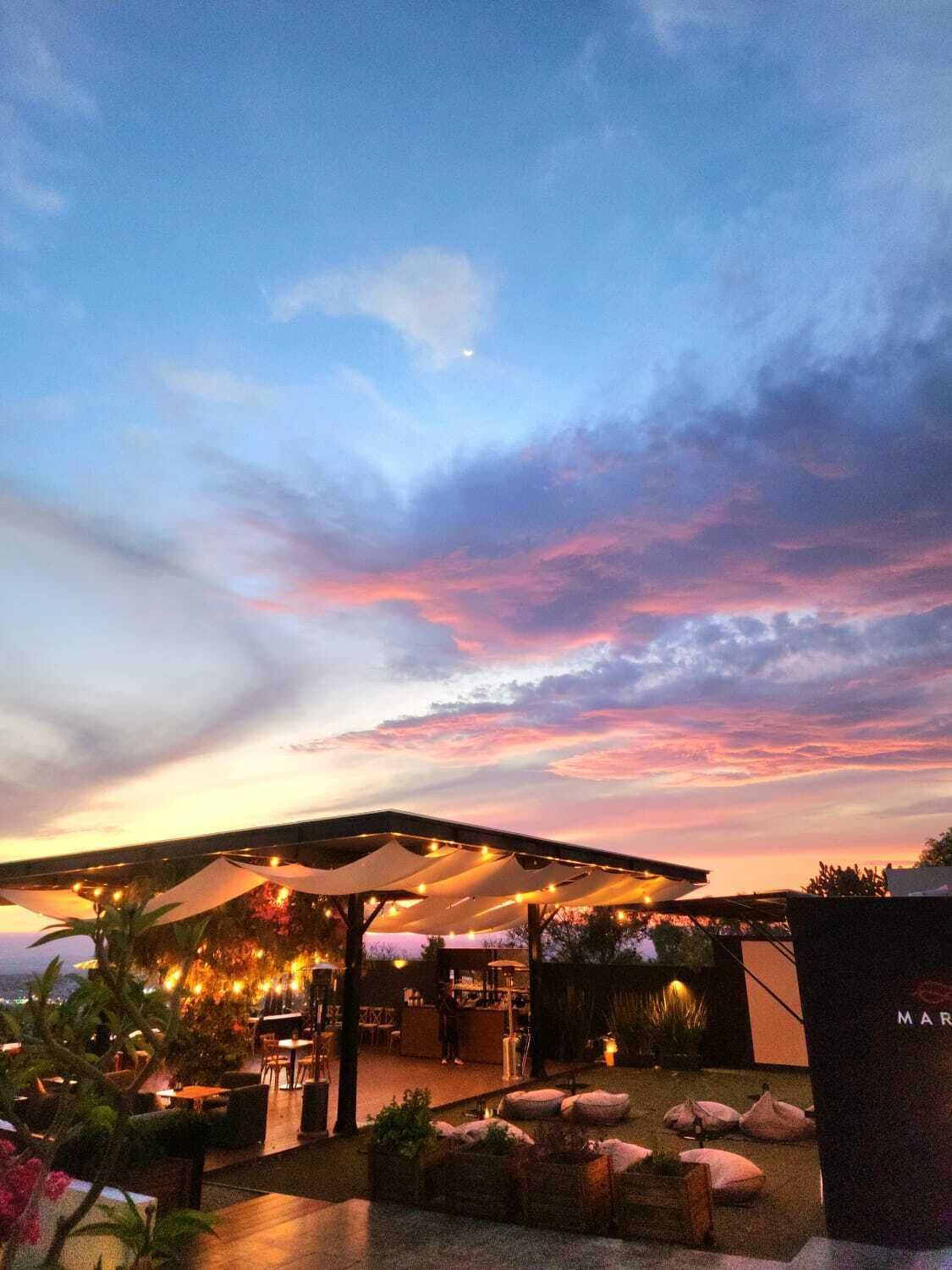 An outdoor seating area with umbrellas and string lights, surrounded by plants and a view of the sky.
