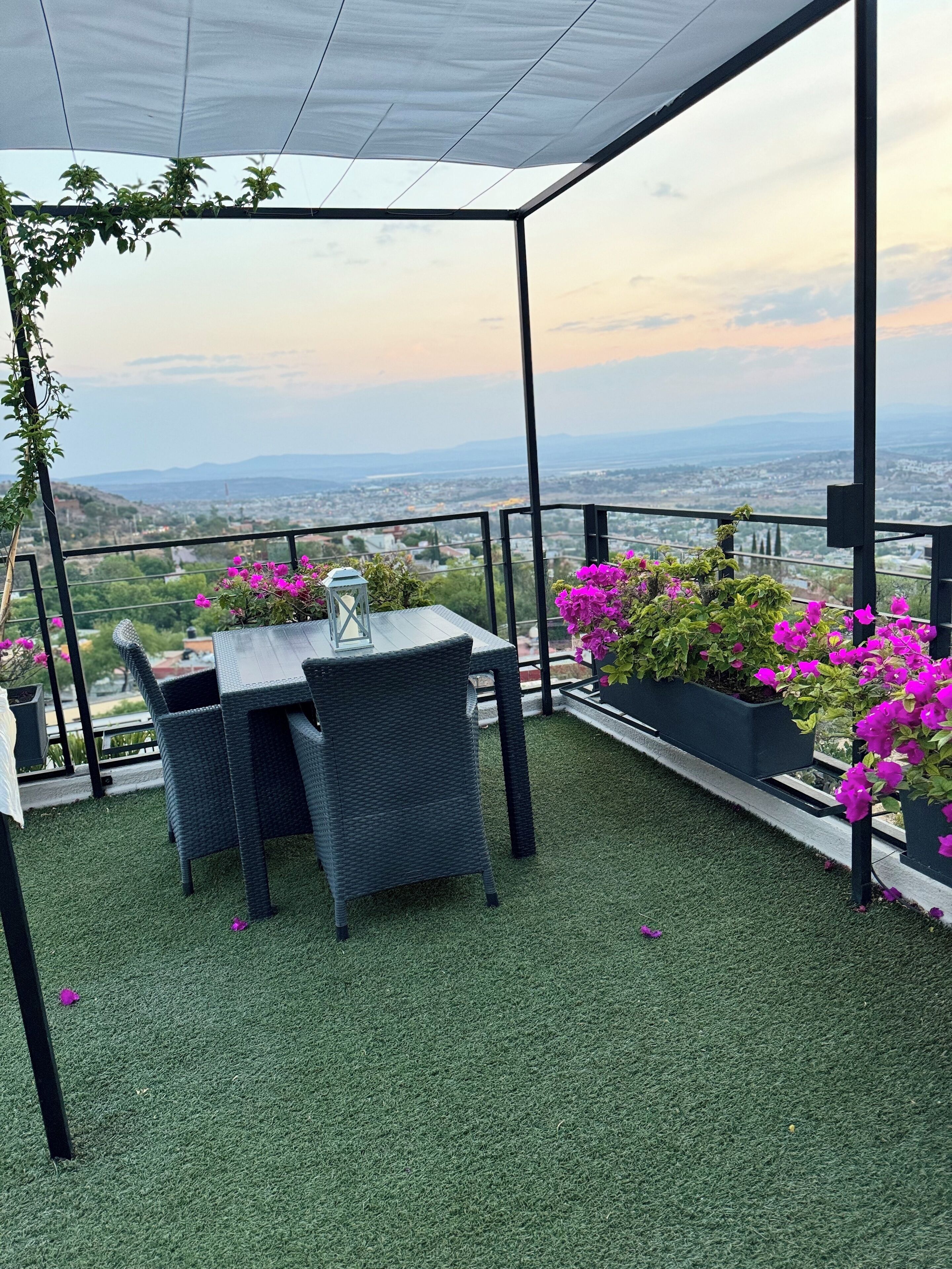 A covered outdoor seating area with a table and chairs, surrounded by artificial grass and potted plants with pink flowers.