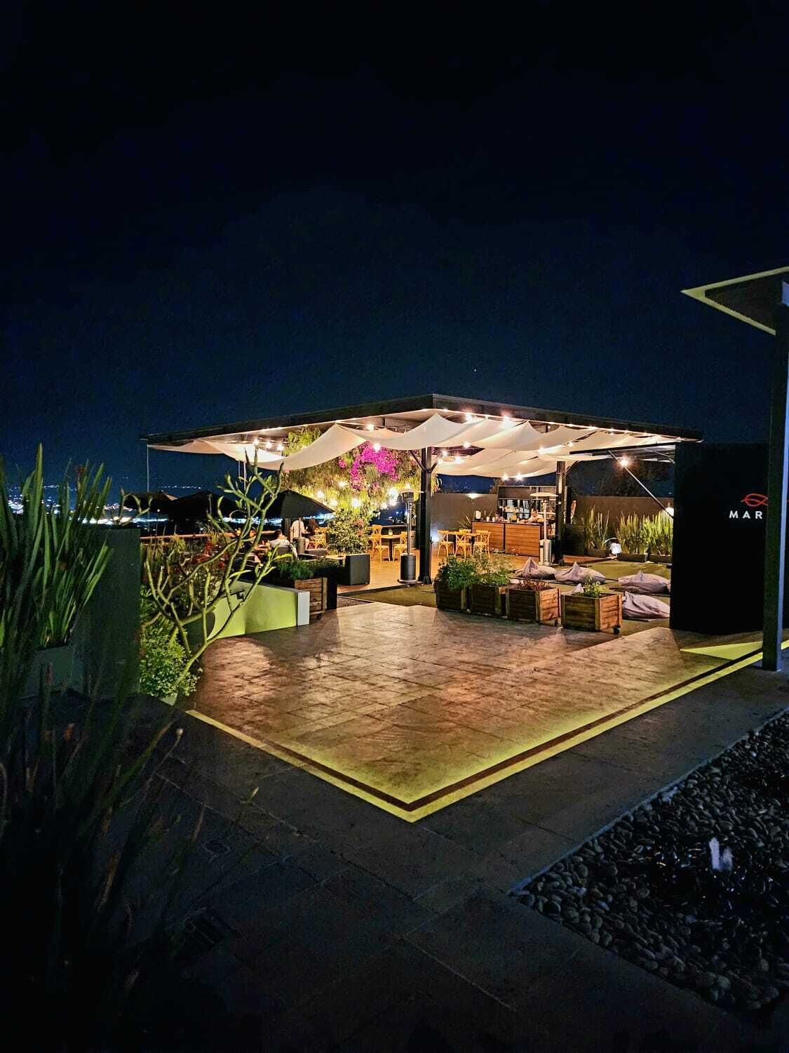 An outdoor patio area with seating and a bar, surrounded by plants and a water feature.