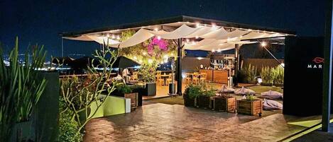An outdoor patio area with seating and a bar, surrounded by plants and a water feature.