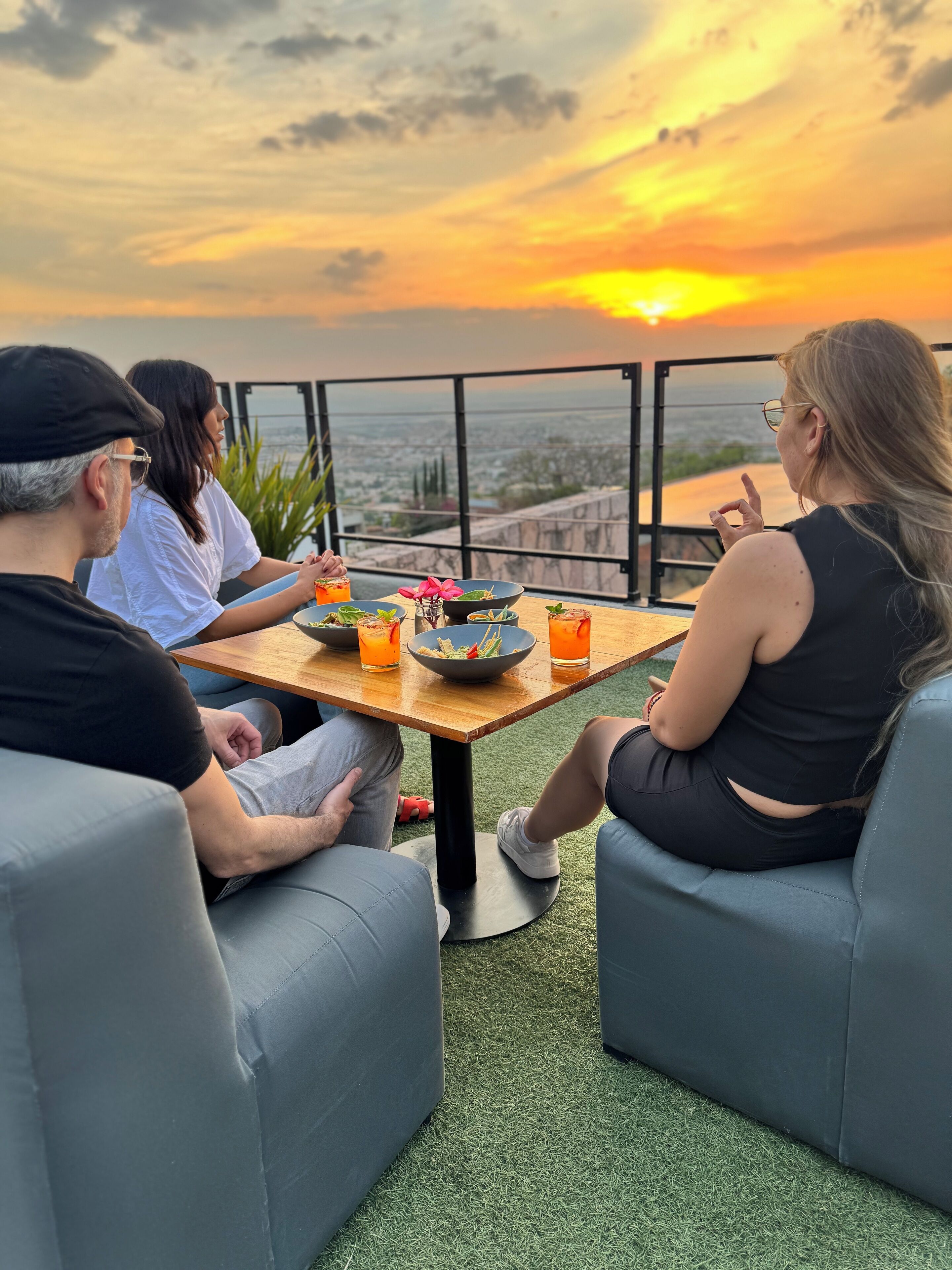 A group of people seated at a table with drinks and a bowl of salad, overlooking a scenic sunset view.