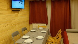 Une pièce aux murs lambrissés, avec une table à manger dressée pour six personnes, un téléviseur fixé au mur et un coin salon confortable agrémenté de rideaux rouges.