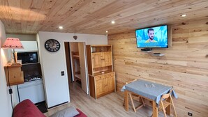Une pièce chaleureuse avec un plafond en bois, un téléviseur fixé au mur, une petite table recouverte d’une nappe et un coin cuisine équipé d’une cuisinière et de placards.